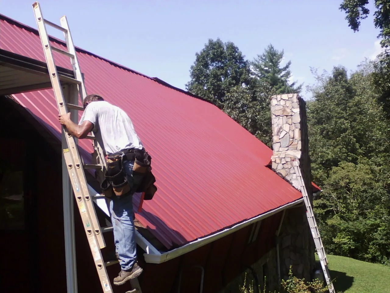 Roofer on a ladder beside a red metal roof and stone chimney — Soffit & Fascia Repair by Roofing Brothers in Willow Hill